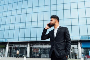 A modern Indian man is talking on the phone, he is in a black suit, behind him a large building, a busy office worker