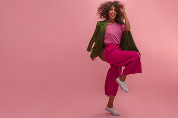 Full size shot of african model jumping and touching her afro haircut happily. Enjoying natural make up with red lipstick and vintage green jacket on the pink costume, standing in front of rosy wall.