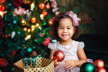 Fototapeta na wymiar Portrait of a little African girl on the background of a decorated Christmas tree. The child is holding a ball in his hands.