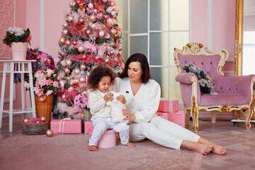 A Caucasian mother and a mixed-race daughter in white sweaters are sitting in a room near a decorated Christmas tree with gifts.