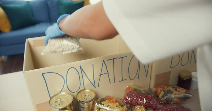Young Asia teenage girl volunteer wear gloves packing food and drinks donation into donations paper boxes while working in charitable foundation. Social worker, food bank and coronavirus concept.