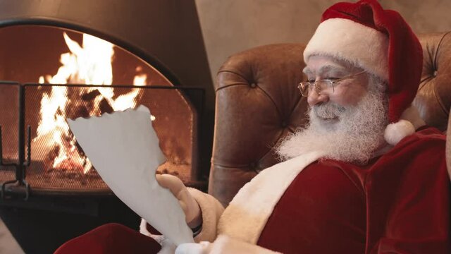Low angle of happy Santa Claus wearing red and white costume with hat, sitting in armchair by fireplace at home, reading letter, then smiling posing on camera