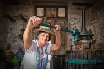 Grape harvest: Old woman smiling winemaker  working on a traditional winepress for the must pressing. Old winery background