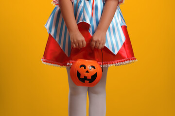 Little girl in a carnival costume for Halloween on a yellow background. The kid is holding a bucket with a pumpkin lantern.