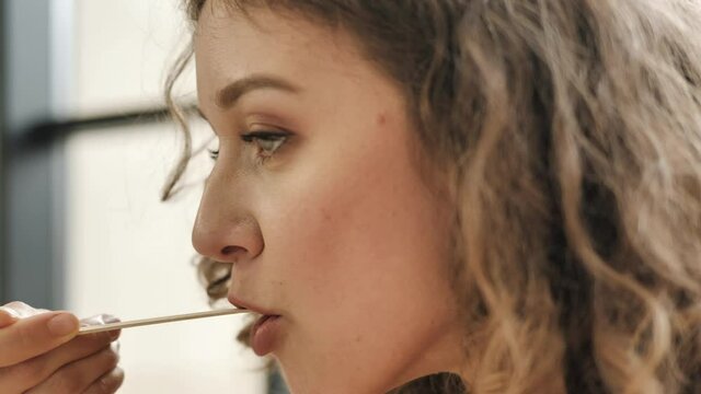 Curly haired young woman with light makeup licks lips eating dish with brown wooden fork and chews up food slowly extreme closeup
