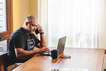 Hombre latino de edad media trabajando desde casa utilizando un celular y computadora