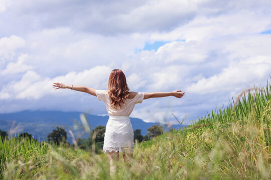A young woman raises both her hands above her head to invoke God's blessing with the faith and power she has for God on the blurry background of a green rice field in the morning.