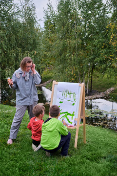 Mother teaches to paint with two boys pupils. Woman teacher artist paints with children on paper nature and trees by the river