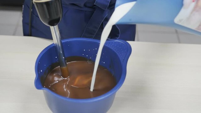 A woman pastry chef with one hand pours cream into hot chocolate, with the other hand simultaneously whips the resulting mass in a blue bowl with a blender. Slow motion