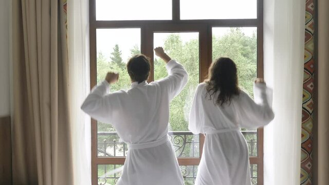 Happy young man and curly haired woman newlyweds in bathrobes dance clap hands and kiss standing near balcony against nature closeup