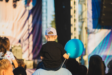 A boy in a black t-shirt with a blue balloon sits on his dad's shoulders at a concert