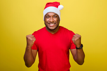 Cheerful young African-American Santa man in a red T-shirt and a Christmas hat makes a winner's gesture, celebrates with clenched fists and says YES, isolated on a yellow background studio portrait