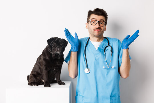 Clueless Doctor Veteriantian In Scrubs Shrugging, Staring At Camera Confused While Dog Sitting On Examination Table And Waiting For Check-up, White Background