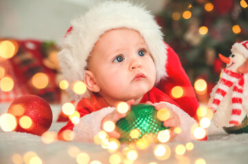 Baby boy dressed as Santa Claus for Christmas plays with wooden train and Christmas decorations in living room...