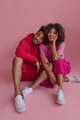 Image of buddies sitting on floor in photo studio pinkish background and look camera. Portrait two latin best friends in trendy outfits showing gladness and enjoying moments.