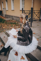 Blonde ballerina in white dress and black jacket reading a book while sitting on the steps of the street in autumn © Alex Barera