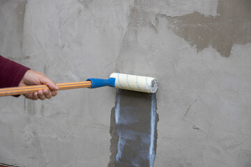 A worker applies the primer to the wall with a long-handled roller. A man's hand rolls a primer...