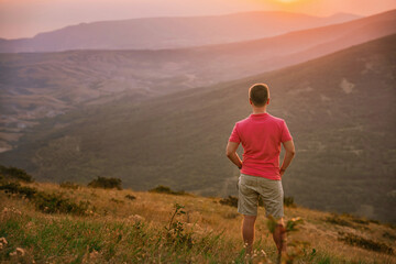 Fototapeta premium A young blond man in a pink shirt conquered the top of the mountain at sunset. Beautiful mountain landscape with a gradient. The concept of enjoying nature.