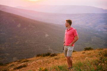 Fototapeta premium A young blond man in a pink shirt conquered the top of the mountain at sunset. Beautiful mountain landscape with a gradient. The concept of enjoying nature.
