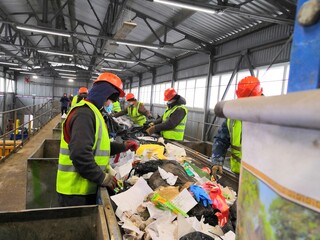 Fototapeta premium male migrant workers in green vests and orange helmets are sorting household garbage on the automatic tape of the garbage landfill