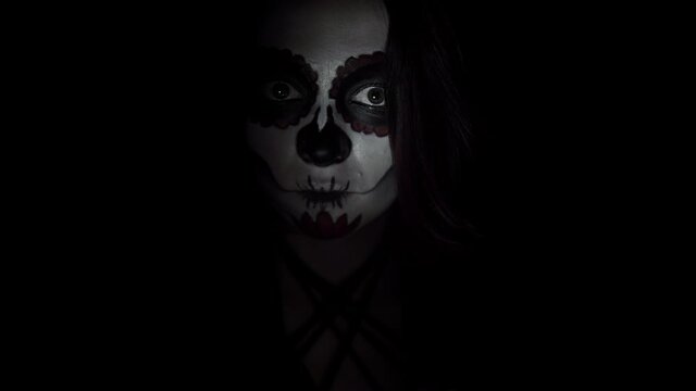 Scary woman with santa muerte make up in close-up on black background is illuminated by a flashlight. A lady with painted face in close-up looks steadily and bends slowly her head on black background.