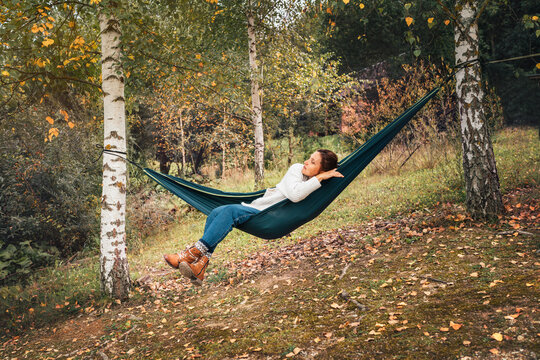  Young woman enjoying warm autumn weather and relaxing in swinging hammock between the birch trees. Out-of-town Outdoor Recreation in Nature concept image.