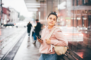 Young woman with glasses and mobile phone waits for bus on public transport stop against large city blurry street in rainy evening side view