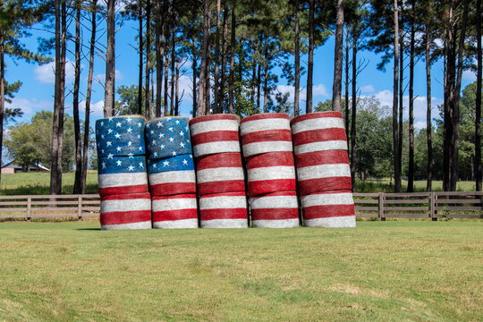 American flag painted on hay bales in a farm field against a backdrop of pine trees and blue sky. Patriotic theme.