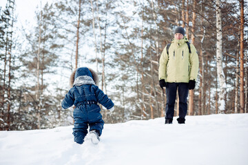 Fototapeta na wymiar Little son happily going towards his dad. Father and baby playing in winter forest outdoors. Man with backpack and toddler boy in blue overalls walking in snowy nature park.