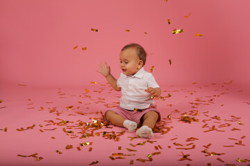 a beautiful happy little boy in a white T-shirt and red shorts is sitting on a pink confetti background