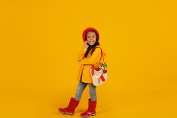 a girl in a red beret with pink glasses in a yellow raincoat and red boots has a bag with autumn flowers on her shoulder