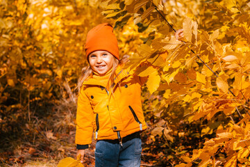 A cheerful little girl in the autumn in a yellow park jacket walks in the autumn park