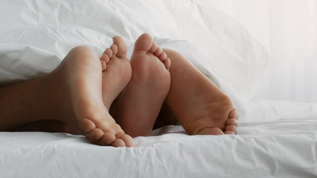 Close up shot unrecognizable diverse couple feets under blanket, partners making love in bed, slow motion