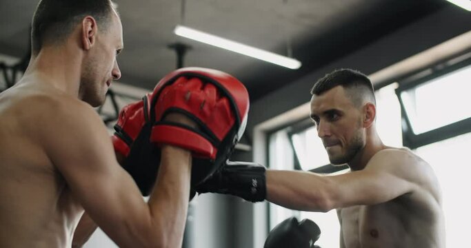 Low angle camera: fighter in boxer gloves is practising his strikes with trainer in slow motion in the gym, kickboxers are training, 4k 120fps Prores HQ