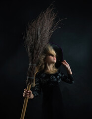 Little girl on a dark background in a witch costume and a wide-brimmed hat with a broom for Halloween.