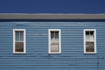 Full frame partial view of a blue painted old wooden building facade with three white windows under deep blue sky