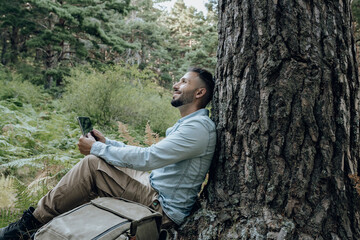 Thoughtful man holding digital tablet while leaning on tree trunk by backpack