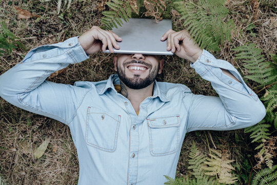 Carefree man lying on plants while covering face with digital tablet in forest