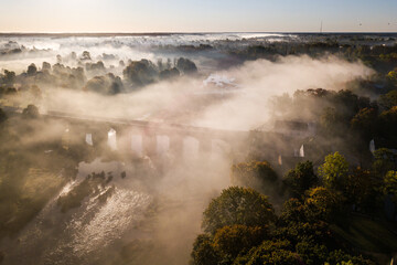 Venta Rapid waterfall, the widest waterfall in Europe and long red brick bridge in foggy, sunny autumn morning, Kuldiga, Latvia. Captured from above.