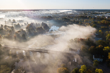 Venta Rapid waterfall, the widest waterfall in Europe and long red brick bridge in foggy, sunny autumn morning, Kuldiga, Latvia. Captured from above.