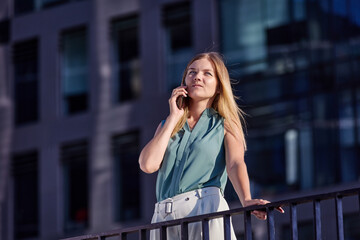 European woman is talking by smartphone near office building.