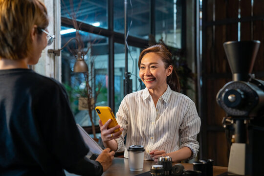 Asian Woman Customer Using Smartphone Scanning Bar Code Make Contactless Payment To Barsita At Cashier Counter In Coffee Shop. Small Business With Modern Mobile Banking Electronic Payment Concept