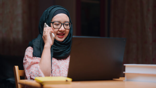 An office worker for an Asian Muslim woman sitting in front of a laptop computer at her desk and talking on a mobile phone and working at the office.