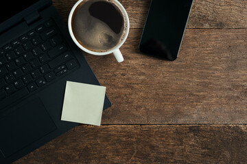 Laptop with empty note paper sheet coffee cup smartphone on wooden office table, Top view with copy space.