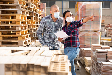 Woman manager in face mask discussing order list with man worker at warehouse of building materials