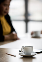 Close up shot of white hot coffee cup and small dish on meeting table near pencil while unidentified unrecognizable businessperson take short break after discussing and planning in blurred background