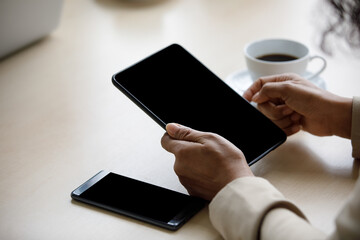 Close up shot of blank black screen touchscreen tablet computer holding in hands of unrecognizable unidentified businesswoman for advertising text and copy space near smartphone and hot coffee cup