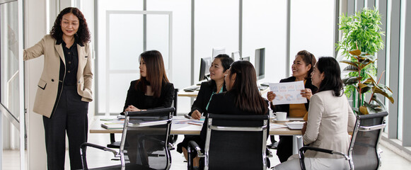 Asian middle aged female businesswoman in casual suit standing smiling presenting pointing information on glass board while colleagues sitting at meeting table clapping hands in conference room