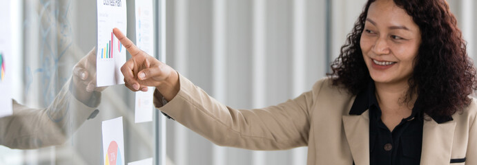Asian middle aged female curly hair businesswoman in casual suit standing smiling presenting pointing business plan graph chart diagram paperwork document on glass board in company meeting room