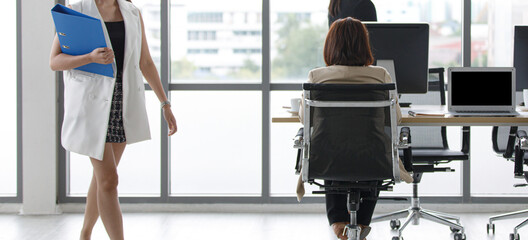 Unidentified unrecognizable female successful businesswoman secretary in formal business suit sitting on chair working typing at office table while other officer staffs holding folder walking around